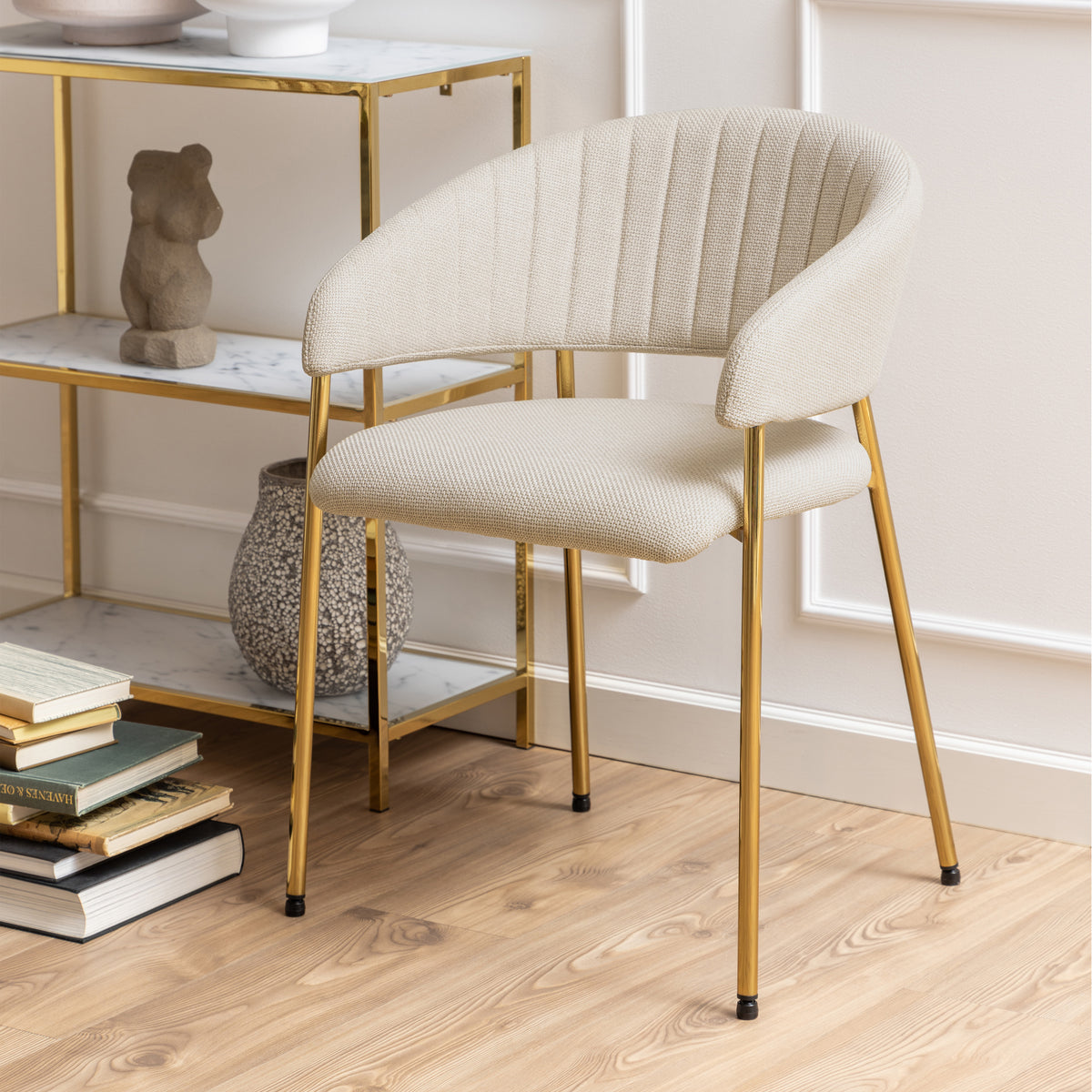 Sedia Brooke beige con gambe dorate in un ambiente moderno, accanto a una libreria decorativa e pavimento in legno.