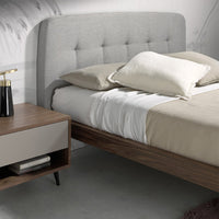 Dettaglio del Letto Luxor con testiera capitonné in tessuto grigio e struttura in legno noce, vicino a un comodino.
