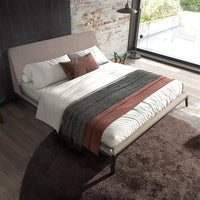 Letto Mara matrimoniale in tessuto grigio con cuscini e coperta, ambientazione moderna con luce naturale e tappeto marrone.